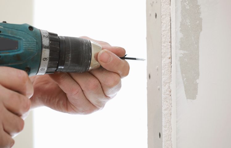 The installer worker in safe medical mask and overalls fixes a detail of the stiffener element with screws using an electric screwdriver to the wall of the metal structure.